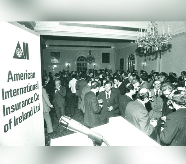 People in a room and a sign with "American International Insurance Co Ireland Ltd" written on it in 1976
